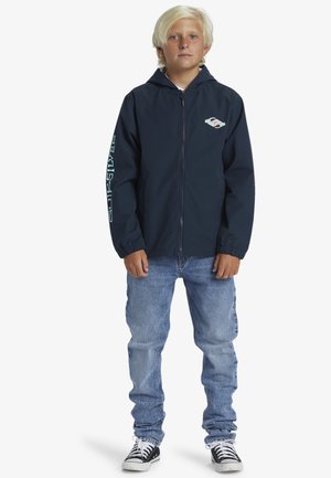 Marinblå zip-up jacka med huva, elastiska ärmslut och en vit logotyp på bröstet. Kombinerad med ljusblå jeans och svart/vita sneakers.