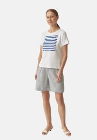 T-shirt en coton blanc avec des rayures horizontales bleues ; associé à un short à rayures grises et blanches. Les chaussures sont des sandales ouvertes blanches.