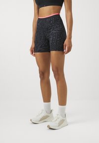 Female wearing black patterned athletic shorts with coral waistband, white socks, and white running shoes standing against a plain background.