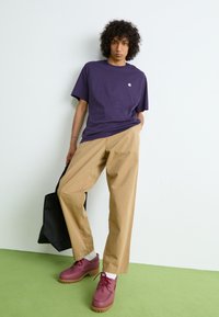 Purple cotton t-shirt, beige wide-leg trousers, and burgundy shoes with a textured sole. Black tote bag added for detail. Green floor surface.