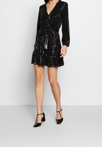 Robe noire à sequins avec un décolleté en V, des manches longues, une taille nouée et un ourlet froncé. Assortie à des talons en vernis noir. Texture lisse.