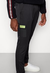 Černé sportovní kalhoty s texturovaným povrchem, elastickými manžetami na kotnících a zelenou náprsní kapsou s logem značky na levém stehně.