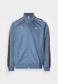 Chaqueta deportiva azul con detalles en blanco, cierre de cremallera en el frente y cuello alto. Presenta logotipos en el pecho y bolsillos laterales. Textura de tela suave.