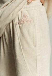 Beige joggers met een zachte textuur, voorzien van een roze geborduurd logo op de zak en een roze merkband langs de zijkant.