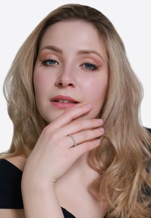 Pierścionek z białego złota z wieloma małymi przezroczystymi kamieniami, ustawiony na dłoni opierającej się o twarz. Modelka ma długie, falujące blond włosy i subtelny makijaż.