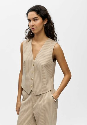 Frau mit langen lockigen Haaren, die eine beige, gestreifte ärmellose Weste und passende Hose trägt, steht mit einer Hand in der Tasche vor weißem Hintergrund.