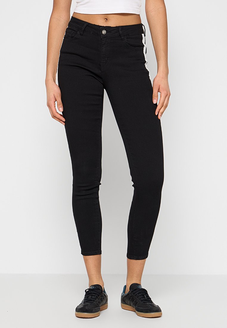 Jean skinny en denim noir à taille haute, avec une fermeture à bouton, des poches avant et des bandes latérales, associés à des chaussures de sport noires.