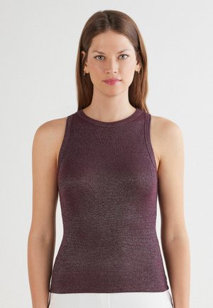 Vrouw met stijlbruin haar in een mouwloze, getailleerde, bordeauxrode glinsterende top tegen een effen witte achtergrond.