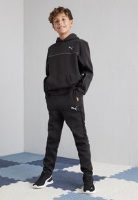 Conjunto deportivo negro con sudadera con capucha y pantalones ajustados. Presenta detalles de piping blanco y un pequeño logo. Tejido texturizado y ajuste ceñido.