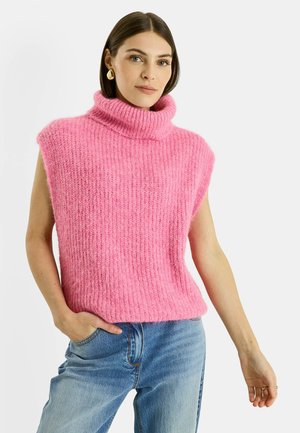 Mujer con cabello castaño liso, que lleva pendientes dorados en forma de lágrima, un suéter de cuello alto sin mangas de punto rosa y jeans azules, con una mano en el bolsillo.