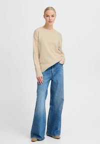 Maglione beige con maniche lunghe, scollatura rotonda, abbinato a jeans svasati azzurri chiari. Tessuto liscio con polsini a costine, vestibilità casual.
