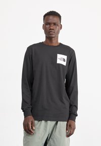 Schwarzes langärmliges T-Shirt aus Baumwolle mit einem weißen Logo auf der Brusttaschenregion. Texturierte Bündchen und eine lockere Passform.