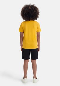 Παιδί με σγουρά μαλλιά στέκεται με την πλάτη προς τα εμπρός, φοράει κίτρινο κοντομάνικο πουκάμισο, μαύρο σορτς και λευκά αθλητικά παπούτσια σε λευκό φόντο.