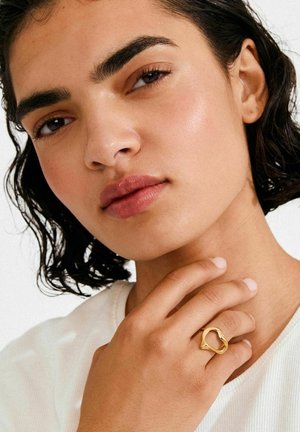 Gouden ring met abstracte vorm, gedragen aan vingers, met een gladde afwerking en open ontwerp. Hand tegen een neutrale achtergrond.