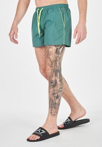 Boxer da bagno verdi con cordino e accenti gialli, abbinati a ciabatte nere con logo. Sono visibili notevoli tatuaggi sulle gambe.