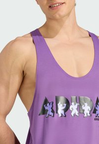 Lilla tanktop med brede stropper, lavet af glat stof med et blankt grafisk print af figurer og mærkenavnet på forsiden.