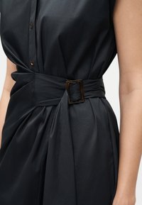 Μαύρο αμάνικο φόρεμα με κουμπιά μπροστά και φαρδύ ζώνη στερεωμένη με σκουρόχρωμη ορθογώνια αγκράφα στη μέση.