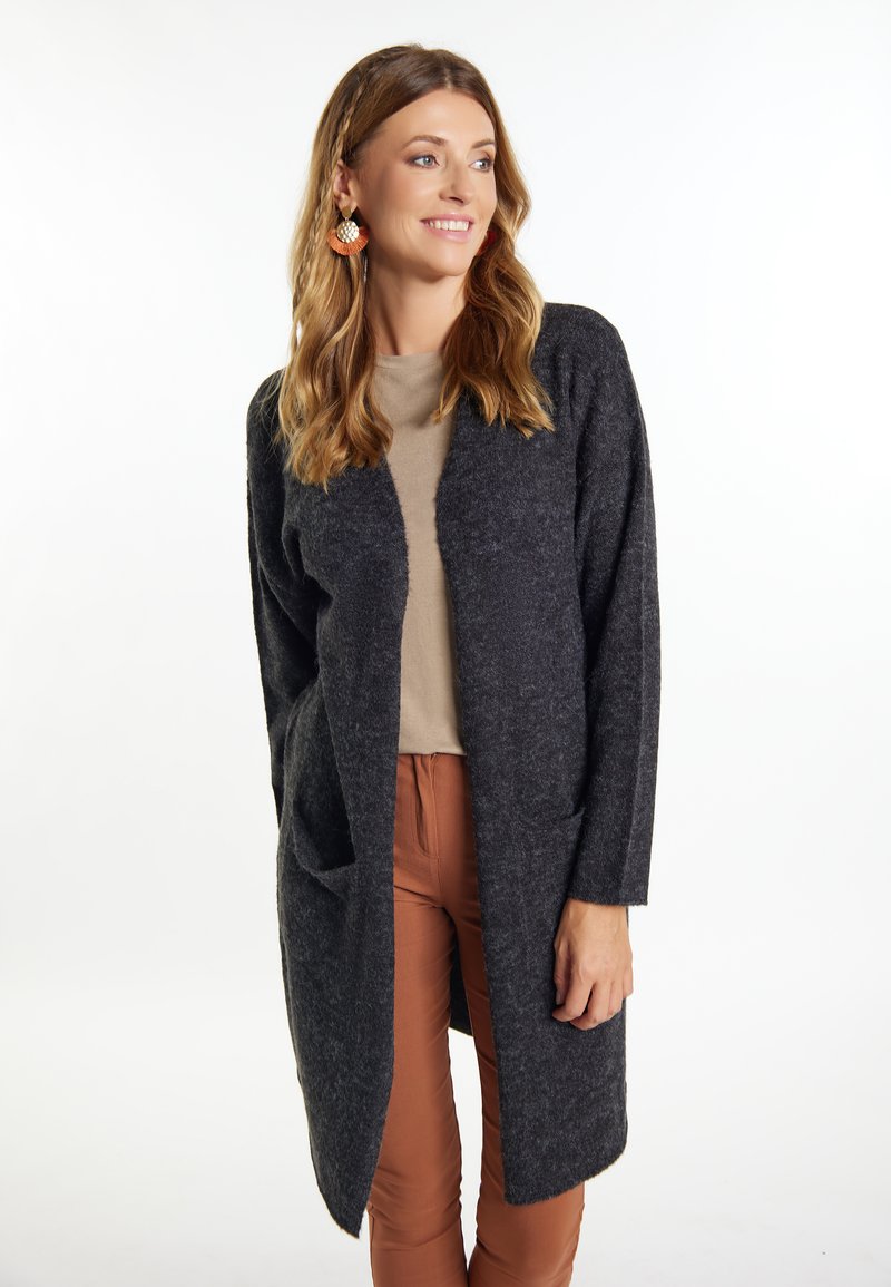 Cardigan lungo grigio scuro realizzato in tessuto testurizzato, con apertura frontale e due tasche frontali, abbinato a un top e pantaloni marrone chiaro.