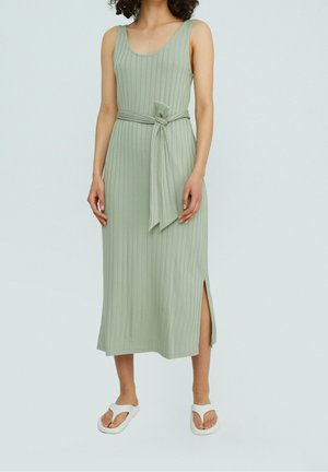 Femme portant une robe midi sans manches côtelée vert clair avec une ceinture nouée et une fente latérale, associée à des sandales blanches.