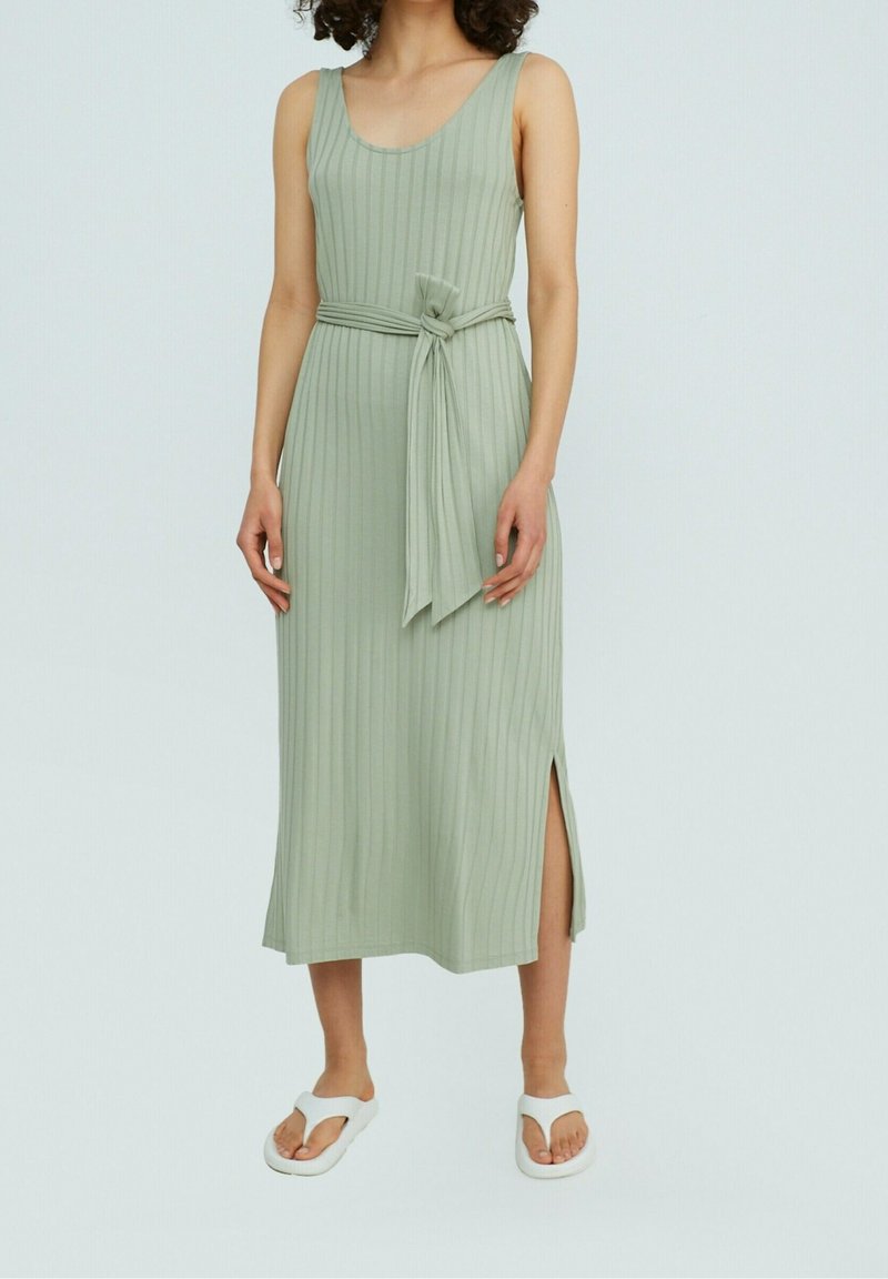 Femme portant une robe midi sans manches côtelée vert clair avec une ceinture nouée et une fente latérale, associée à des sandales blanches.