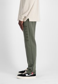 olijfgroene joggingsbroek met reliëftekst "ALPHA INDUSTRIES", gecombineerd met zwart-witte sneakers en een beige langwerpige top.