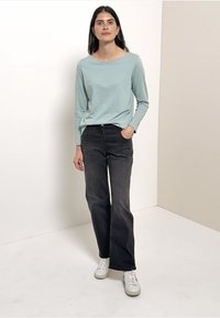 Lysblå langærmet top, afslappet pasform; parret med mørkegrå vide bukser. Modellen bærer hvide sneakers og står foran en neutral baggrund.