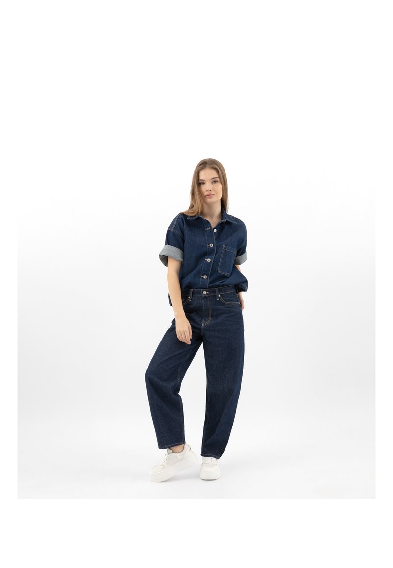 Femme debout portant une chemise en denim bleu foncé à manches courtes retroussées, un jean large assorti et des baskets blanches sur fond blanc.