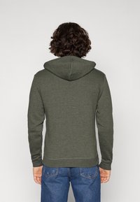 Persona con capelli ricci che indossa una felpa verde oliva e jeans blu, girata di spalle contro uno sfondo bianco semplice.