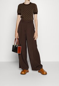 Bruin gebreide korte mouwen trui met geometrisch patroon, gecombineerd met wijde bruine broek. Houdt een tweekleurige handtas met textuuraccenten vast.