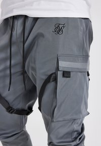 Harmaat työhaalarit, jotka on valmistettu teksturoidusta kankaasta ja joissa on kohokuvioitu logo, sivutaskut ja mustat säädettävät hihnat.