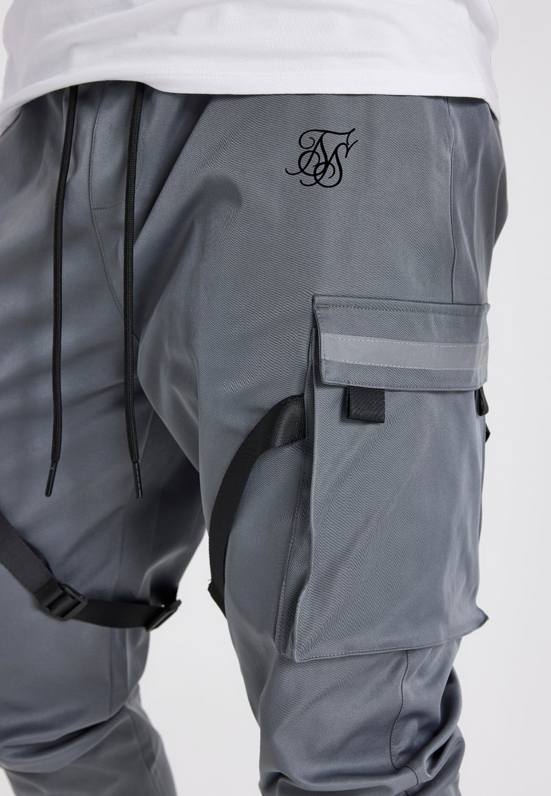 Harmaat työhaalarit, jotka on valmistettu teksturoidusta kankaasta ja joissa on kohokuvioitu logo, sivutaskut ja mustat säädettävät hihnat.