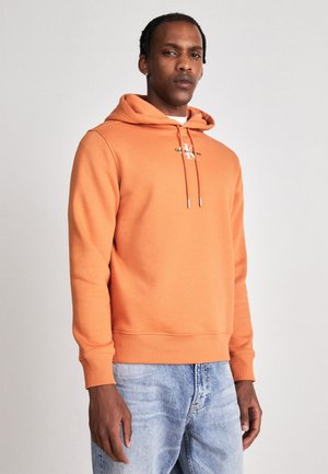 Felpa arancione con cappuccio in morbido misto cotone, caratterizzata da un logo frontale, dettagli con lacci e polsini a coste, abbinata a jeans chiari.