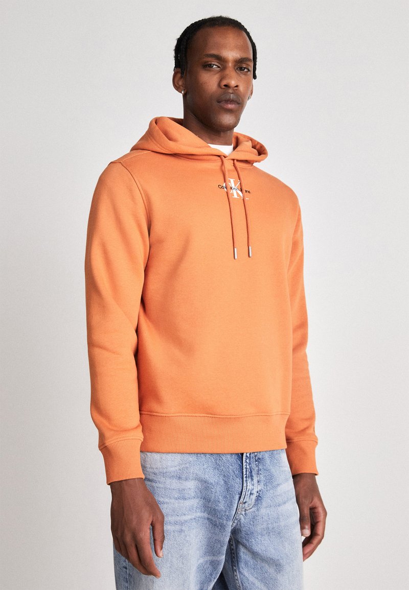 Felpa arancione con cappuccio in morbido misto cotone, caratterizzata da un logo frontale, dettagli con lacci e polsini a coste, abbinata a jeans chiari.
