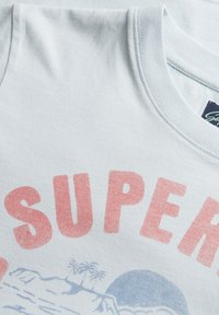 Camiseta de algodón de color azul claro con una gran impresión roja desvanecida con la palabra "SUPER" y un gráfico azul de un paisaje con palmeras.