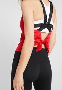 Débardeur de sport rouge avec bretelles noires réglables, doté d'un nœud à l'arrière et d'un design superposé sur un sous-vêtement blanc.