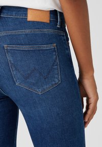 Jeans in denim blu scuro con vita alta, caratterizzati da un'etichetta in pelle marrone sul retro e un distintivo dettaglio di cucitura a forma di triangolo sulla tasca.