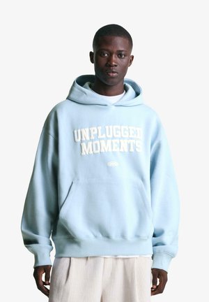 Hellblaue Kapuzenjacke mit "UNPLUGGED MOMENTS" in Weiß, ausgestattet mit einer Fronttasche und gerippten Bündchen, kombiniert mit cremefarbenen gestreiften Hosen.