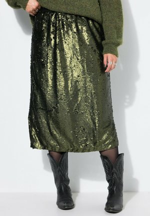 Mujer que lleva una falda brillante de lentejuelas verde oscuro, botas de vaquero negras con estampado y un suéter verde oliva, de pie contra un fondo blanco.