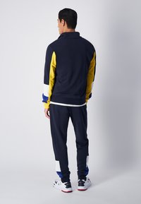 Tuta navy con accenti gialli, bianchi e blu. La giacca ha un colletto alto; i pantaloni hanno una vestibilità affusolata. Materiale testurizzato.