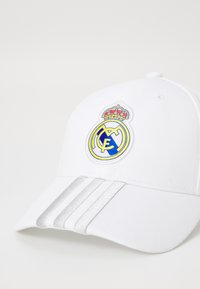 Hvid baseballcap med broderet Real Madrid-logo i gult og blåt samt sølvstriber på skyggen; lavet af struktureret stof.