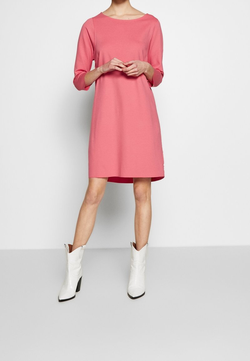 Femme en robe rose longueur genoux avec manches trois-quarts, debout les mains jointes et portant des bottes blanches à talons mi-mollet.