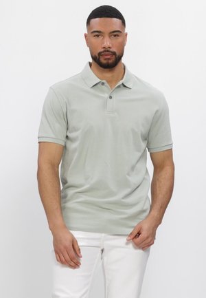 Man met kort zwart haar en baard, gekleed in een lichtgrijs poloshirt en een witte broek, staand tegen een effen witte achtergrond.