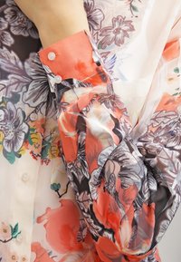Bloemige shirt met een semi-doorzichtige, lichte stof. Voornamelijk rood en zwart met kleurrijke bloempatronen en knoopdetails.