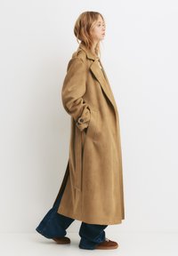 Long manteau en daim beige avec un col à revers, des poches latérales et un devant ouvert, associé à un jean à jambes larges et des baskets marron.