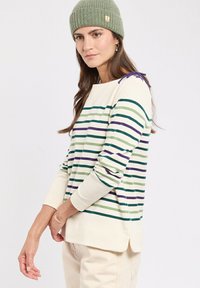 Femme portant un bonnet en tricot vert et un pull crème avec des rayures horizontales vertes, violettes et bleues, se tenant devant un fond blanc.