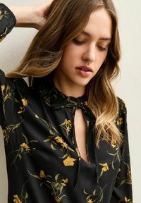 Zwarte blouse met geel bloemenpatroon, voorzien van een strik aan de hals, lange mouwen en een zachte, gladde textuur.