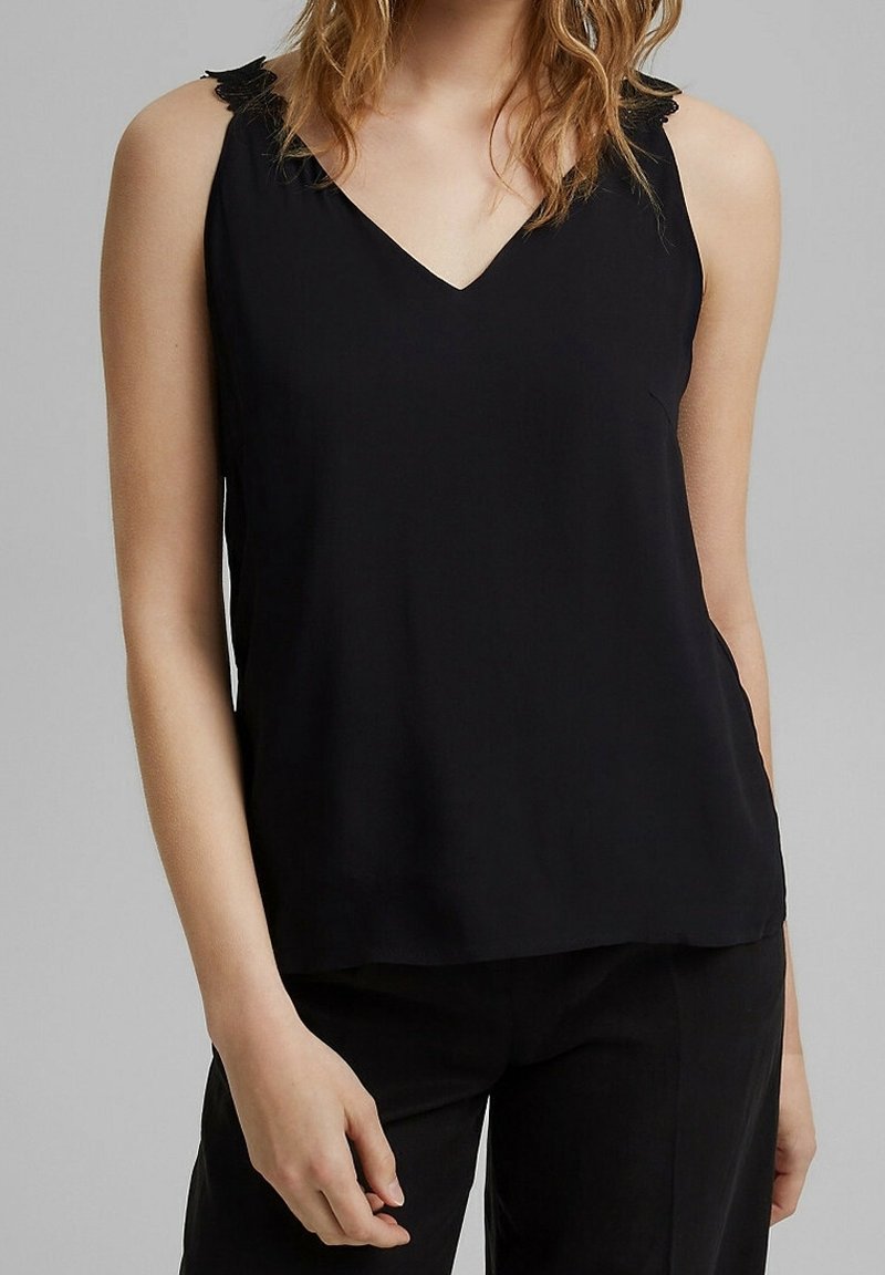 Femme portant un haut noir sans manches avec un col en V et un pantalon noir debout devant un fond gris.
