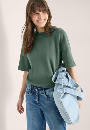Femme souriante portant un haut vert à manches courtes et un jean bleu, tenant un sac fourre-tout en jean bleu clair sur son bras.