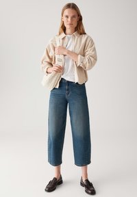 Young woman wearing beige corduroy jacket, white shirt, wide blue jeans, black loafers, holding cream shoulder bag strap.