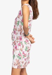 Valkoinen kukkamekko, jossa on vaaleanpunaisia ja vihreitä kuvioita, hihaton malli ja nyöreillä kiristettävä vyötärö. Suora kangas.